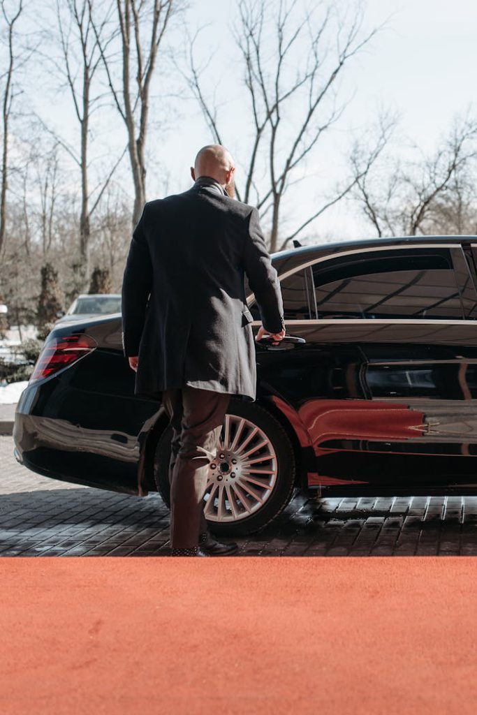 pexels photo 7594130 Bald man in a black suit approaches a luxury black car on a sunny day.